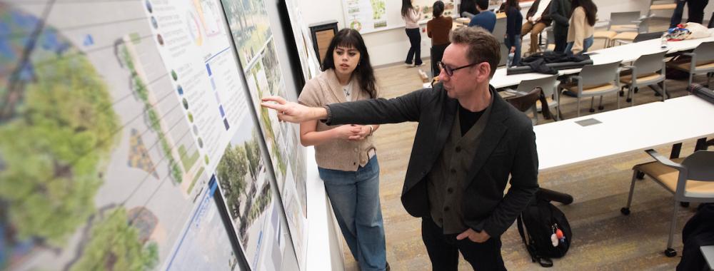 Student and professor stand together near projects posted up on the wall.