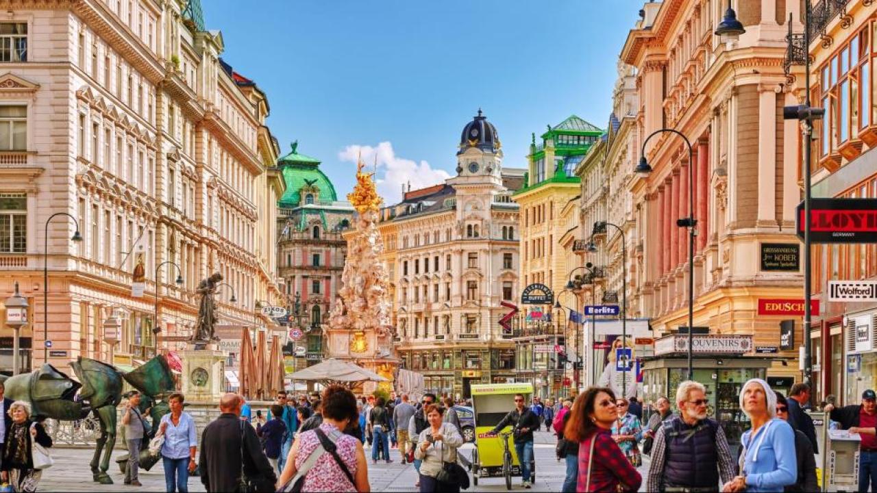 Crowded European pedestrian street lined with ornate historic buildings and green rooftops
