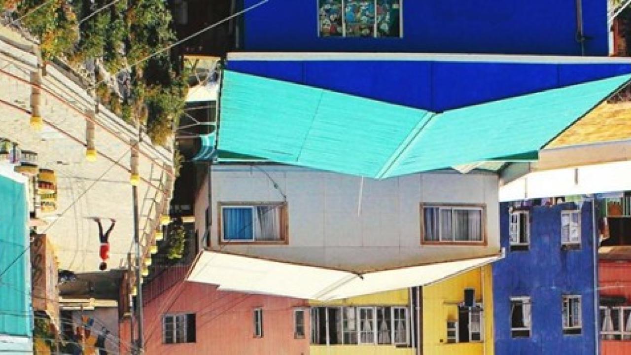 Upside-down image of a colorful urban neighborhood with tightly packed buildings, metal roofs, balconies, and hanging laundry, creating a visually disorienting yet vibrant scene.