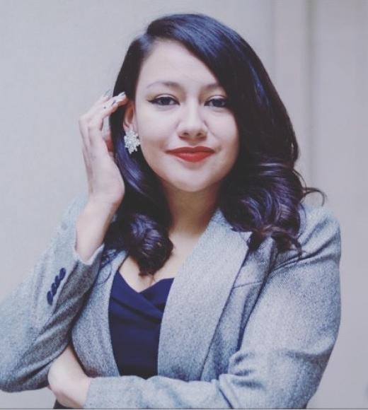 A woman in a grey blazer and dark top stands confidently with one arm crossed and the other hand touching her earring. She has dark wavy hair, red lipstick, and a composed, professional expression.