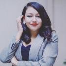A woman in a grey blazer and dark top stands confidently with one arm crossed and the other hand touching her earring. She has dark wavy hair, red lipstick, and a composed, professional expression.