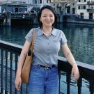 A smiling woman with short dark hair is standing on a bridge next to a body of water. She is wearing a gray polo shirt, blue jeans, and a brown belt with a large silver buckle. She is carrying a brown tote bag on her left shoulder and is leaning on the bridge railing with her right arm. In the background, there is a city with buildings and another bridge.